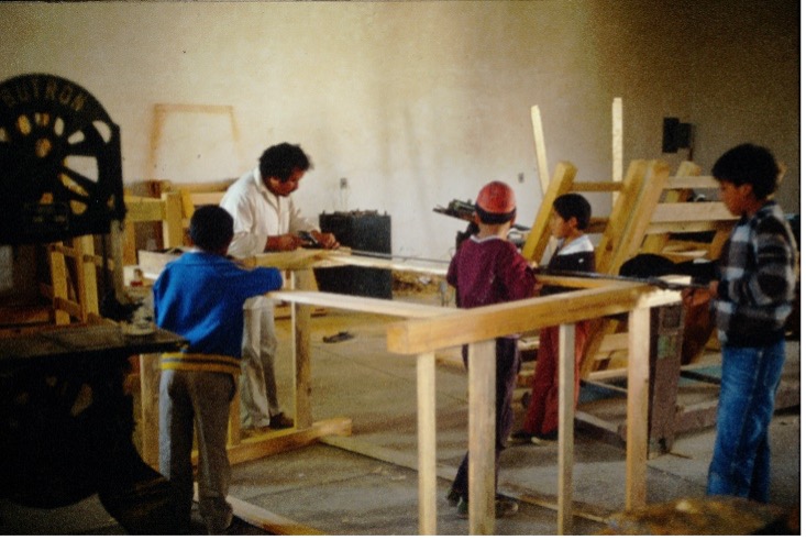 Das Foto zeigt die Holzwerkstatt in einer Kernschule in Atlacomulco in Méxiko. Vier sehr junge männliche Schüler arbeiten zusammen mit ihrem Lehrer am Bau von Etagenbetten. Am linken Bildrand steht eine Sägemaschine, im Hintergrund sind weitere Bettgestelle zu erkennen. 