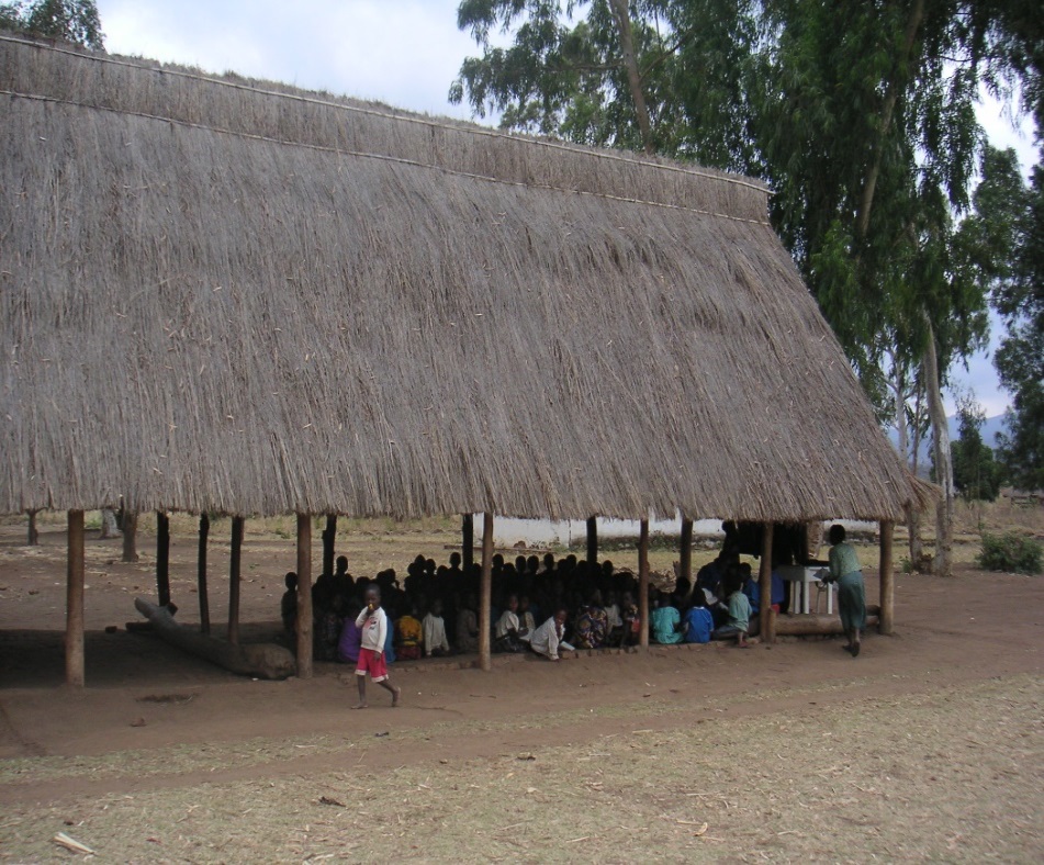 Ein „Schulgebäude“ in Zentralmalawi. Eine hohe Halle aus einfachen Baumstämmen und mit einem strohgedeckten hohen Dach. Etwa dreißig Kinder sitzen auf dem Lehmboden vor einer einfachen Tafel. Im Hintergrund stehen einige grüne Bäume. 