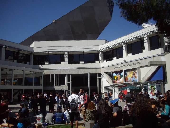 Ein modernes mehrgeschossiges Gebäude aus Beton der San Francisco State University, das in einem Halbkreis gebaut ist und dadurch einen offenen Platz markiert. Im Hintergrund wölbt sich eine kühne Dachkonstruktion in den blauen Himmel, die an ein Segel erinnert. Der Platz ist voll mit Studierenden. In dem Gebäude haben die verschiedenen studentischen Gruppen (für Frauen, für Queers, für Menschen mit Behinderungen, für verschiedene ethnische und kulturelle Gruppen) ihre Büros und Veranstaltungsräume.